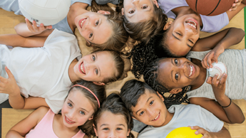 Children lying on the floor looking up smiling holding various sport equipment.
