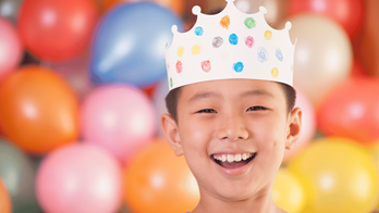 Kid with party hat on smiling with balloons in the background.