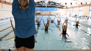 Instructor in a blue swimming top delivering an exercise class at Dawlish Leisure Centre Swimming pool. Several members enjoying, smiling the exercise class whilst in the water.