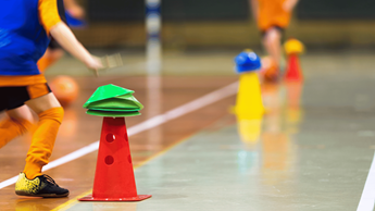 Child in blue bib in a sports hall participating in a junior activity with cones.