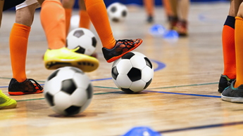 Child in orange socks in a sports hall participating in a junior activity with footballs.